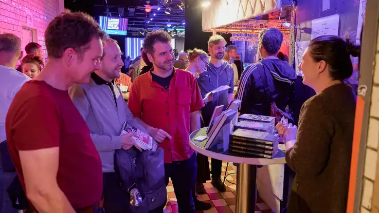 Gruppe von Menschen steht an einem Stehtisch mit gestapelten Büchern und unterhält sich in einem Raum mit Backsteinwand und bunter Beleuchtung.