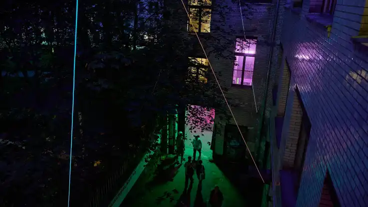 Dunkle Gasse zwischen zwei Häusern mit mehreren Personen, die von bunter Lichtinstallation in Grün, Blau und Violett beleuchtet wird.