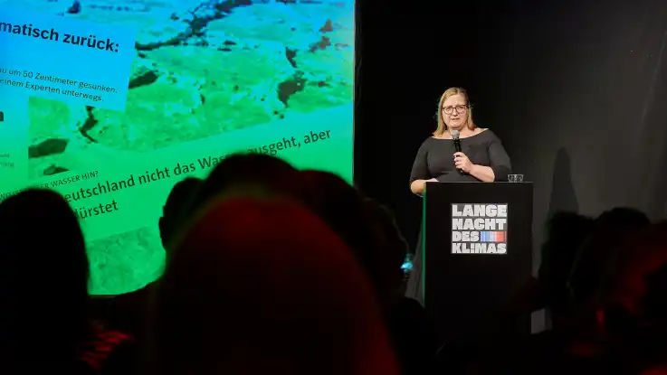 Frau steht mit Mikrofon an einem Rednerpult mit der Aufschrift «Lange Nacht des Klimas» vor einer Leinwand mit Text und Bild, Publikum im Vordergrund.