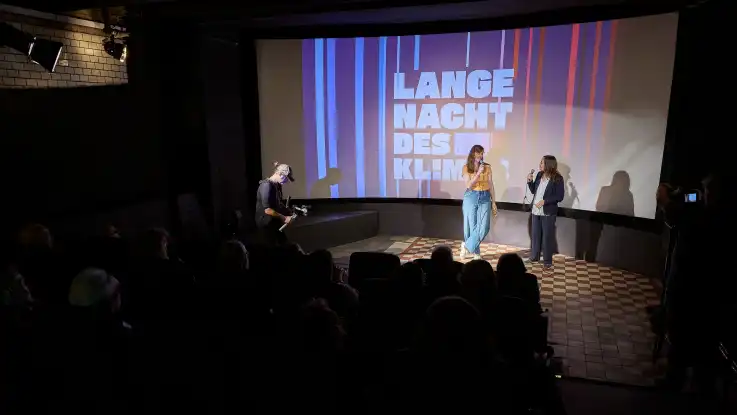 Zwei Frauen stehen auf einer Bühne vor einer Leinwand mit dem Text «Lange Nacht des Klimas» und sprechen in Mikrofone, davor sitzt ein Publikum im dunklen Kinosaal.