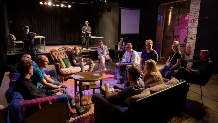 Gruppe von Menschen sitzt in einem Kreis auf Sofas und Stühlen in einem Theater- oder Probenraum mit dunklen Wänden und Teppich auf dem Boden.