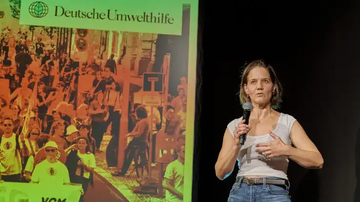 Frau mit Mikrofon vor Leinwand mit Logo der Deutschen Umwelthilfe und Menschenmenge im Hintergrund.
