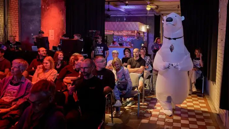 Menschen sitzen in einem Raum mit Kachelfußboden und beobachten eine Person in einem aufblasbaren Eisbärenkostüm, die durch den Gang geht.