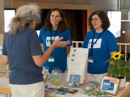 Ein grauhaariger Mann steht an einem kleinen EWS-Infostand und unterhält sich mit zwei EWS-Mitabeiterinnen in blauem T-Shirt mit EWS-Logo.