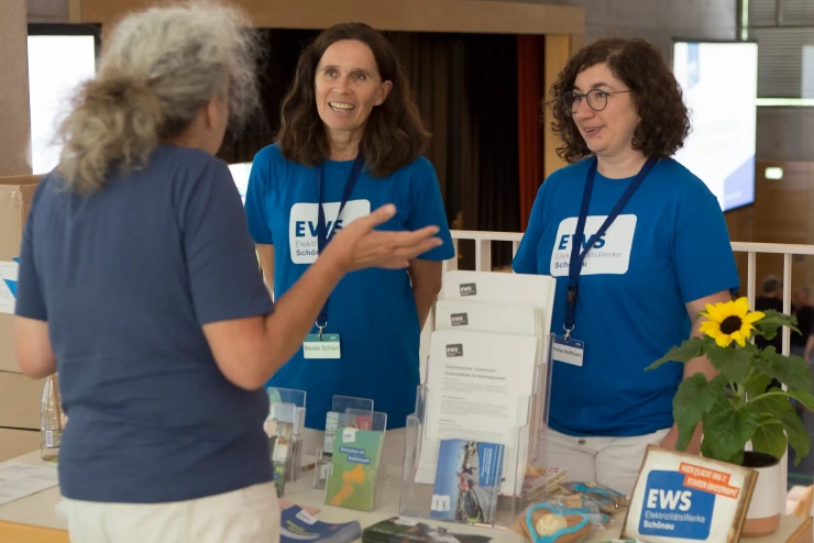 Ein grauhaariger Mann steht an einem kleinen EWS-Infostand und unterhält sich mit zwei EWS-Mitabeiterinnen in blauem T-Shirt mit EWS-Logo.