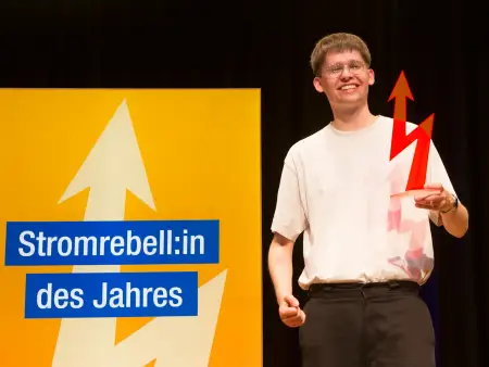 Ein junger Mann in naturweißem T-Shirt ballt die rechte Hand, in der linken hält er eine blitzförmige, rote Trophäe; hinter ihm eine Leuchtstele mit der Aufschrift »Stromrebell:in des Jahres»