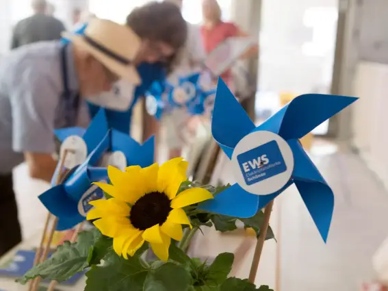 Eine Sonnenblume und kleine, blaue Spielzeugwindräder auf einem Tisch – dahinter sind unscharf Personen zu sehen. 