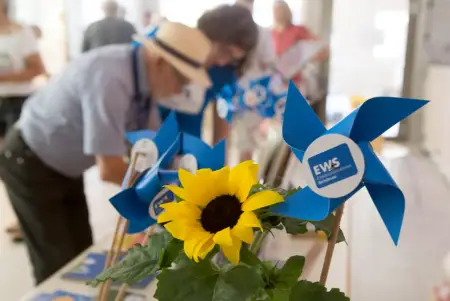 Eine Sonnenblume und kleine, blaue Spielzeugwindräder auf einem Tisch – dahinter sind unscharf Personen zu sehen.